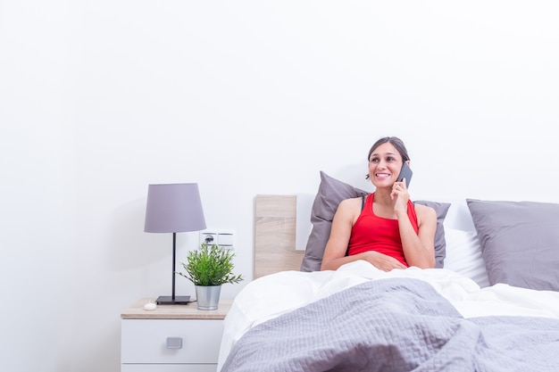 Mujer joven feliz en la cama sonriendo mientras escucha en el teléfono móvil