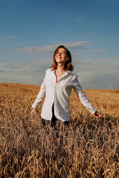 Mujer joven feliz con una camisa blanca en un campo de trigo. Día soleado. Niña sonriendo, concepto de felicidad. Manos a los lados. Foto vertical