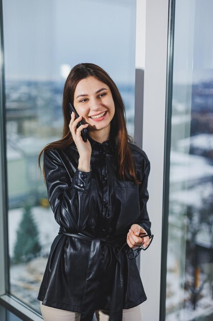 Mujer joven feliz con una camisa negra sonriendo y hablando por teléfono en una oficina moderna con grandes ventanales Trabajo remoto