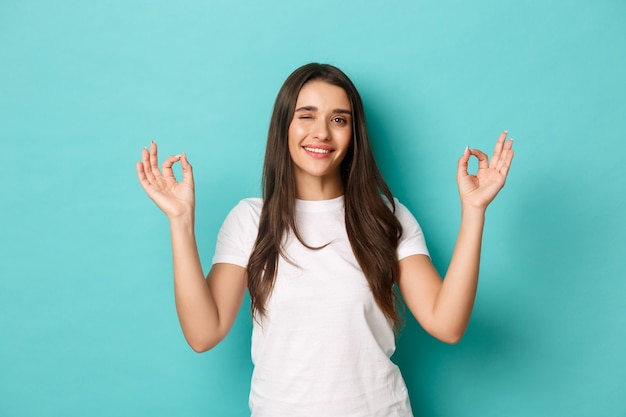 Mujer joven feliz en camiseta blanca, guiñando un ojo y mostrando signos de bien, sonriendo complacido, recomendar