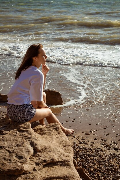 Mujer joven feliz en camiseta blanca y pantalones cortos se sienta en la orilla del mar en verano