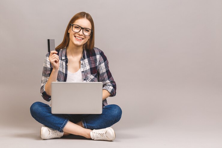 Foto mujer joven feliz en casual sentado en el suelo con ordenador portátil y tarjeta de crédito