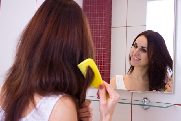 Mujer joven feliz cepillando su largo cabello castaño con peine frente al espejo en el baño