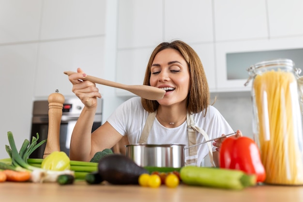 Mujer joven feliz cocinando degustando la cena en una olla de pie en la cocina moderna en casa ama de casa preparando comida saludable sonriendo hogar y nutrición recetas de dieta concepto