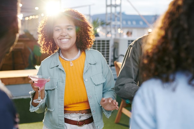 Mujer joven feliz con cóctel mirando a uno de los amigos mientras disfruta de la fiesta