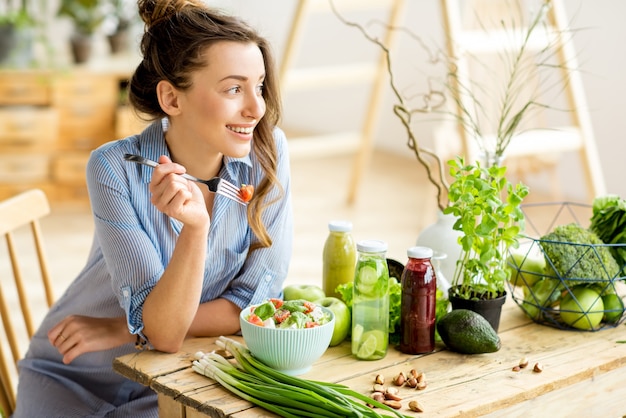 Mujer joven y feliz comiendo ensalada saludable sentado en la mesa con ingredientes frescos verdes en el interior