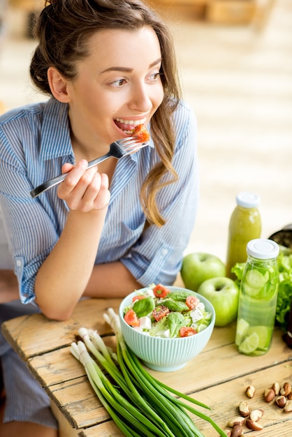 Mujer joven y feliz comiendo ensalada saludable sentado en la mesa con ingredientes frescos verdes en el interior