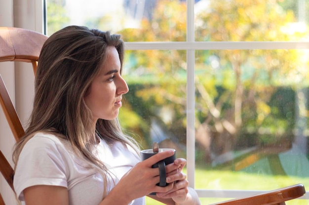 Mujer joven feliz desayunando tomando una taza de café mientras mira desde la ventana al jardín