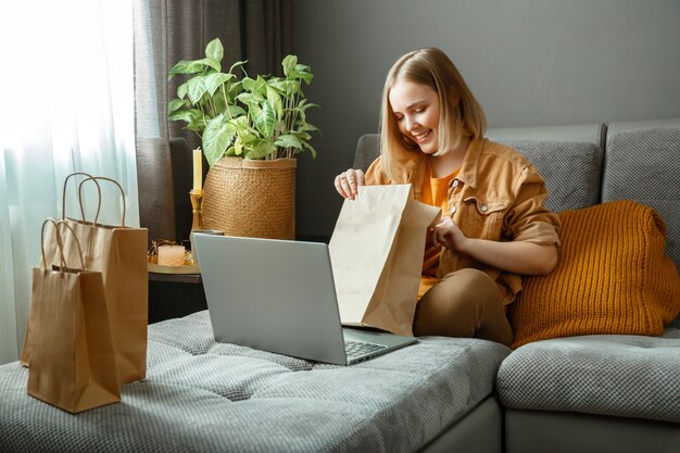 Mujer joven feliz desembalaje de pedidos en línea productos o alimentos compras en línea pedidos de entrega adolescente niña relajarse en el sofá considerando compras con laptop mock up bolsas de papel