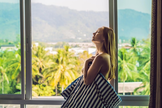 Mujer joven feliz se despertó por la mañana en el dormitorio junto a la ventana con hermosas vistas a la montaña