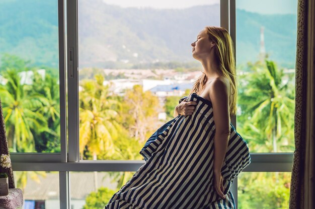 Mujer joven feliz se despertó por la mañana en el dormitorio junto a la ventana con hermosas vistas a la montaña