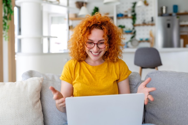 Mujer joven feliz divirtiéndose haciendo videollamadas usando una computadora portátil en su casa saludando con la mano videoconferencia llamando a una computadora portátil sentada en la distancia del sofá aprendiendo zoom en línea reunión virtual en casa