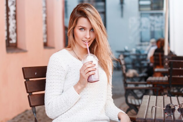 Mujer joven feliz con un elegante suéter de punto está sentada en una silla de madera en un café de verano en la calle en un cálido día de verano y bebiendo un dulce cóctel. Chica alegre bebe una copa.