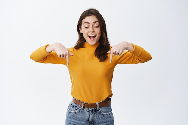 Mujer joven feliz y emocionada que parece hipnotizada y fascinada hacia abajo señalando con el dedo el espacio vacío inferior para el anuncio que muestra el fondo blanco del logotipo