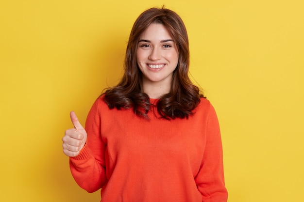 Mujer joven feliz europea con ropa naranja, levanta el dedo, muestra como con el pulgar hacia arriba sobre amarillo, niña expresando positivo, tiene mirada satisfecha.