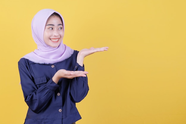 Foto mujer joven feliz con expresión facial feliz muestra algo en su mano vistiendo hijab y ropa casual en amarillo