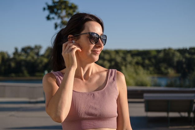 Mujer joven feliz fitness en gafas de sol terminar de correr en la ciudad. Ponte los auriculares. tener un descanso. disfruta escuchando música en auriculares. atardecer. estilo de vida saludable. libertad. entrenamiento al aire libre.