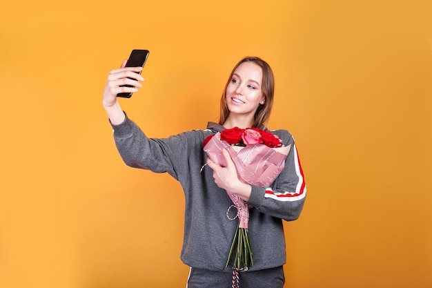 Mujer joven feliz con flores rosas tomando selfie sobre fondo amarillo