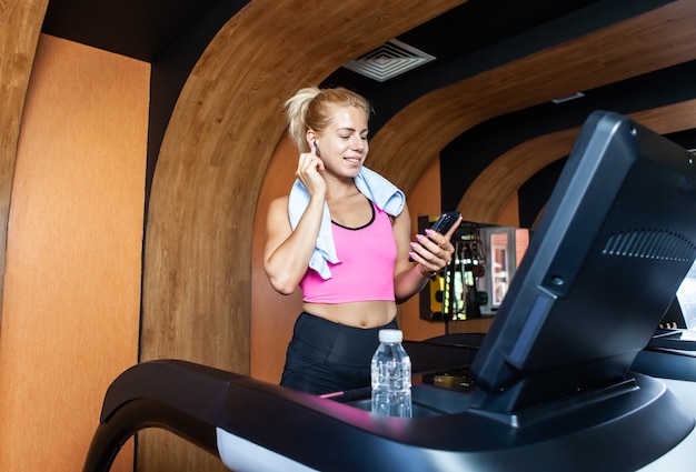 Mujer joven feliz en forma con una toalla sobre los hombros en la caminadora escuchar música en el gimnasio