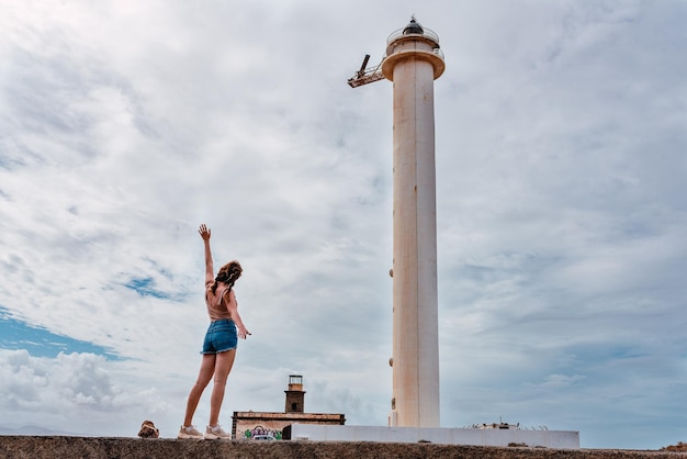 Mujer joven feliz frente a un faro con el brazo extendido haciendo un gesto de libertad