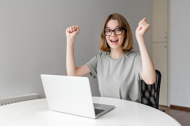 Foto mujer joven feliz con gafas ganando el premio sentado con el portátil en el apartamento