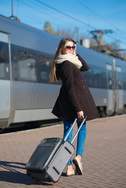 Mujer joven feliz con gafas vestida con abrigo con una gran bolsa de viaje en la estación de tren en el fondo del tren