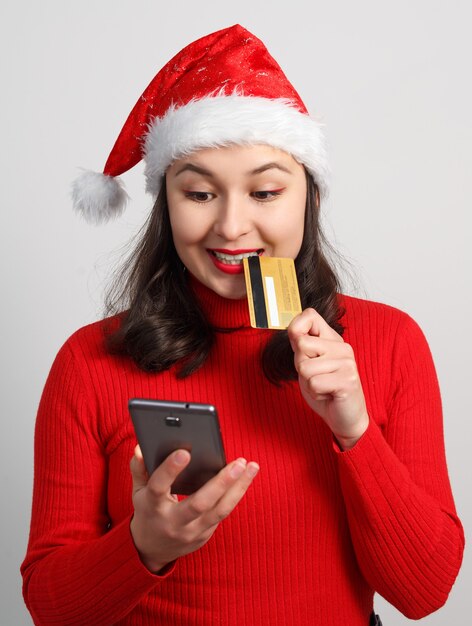 Mujer joven feliz con un gorro de Navidad y un suéter rojo tiene un teléfono inteligente y una tarjeta de crédito en blanco.