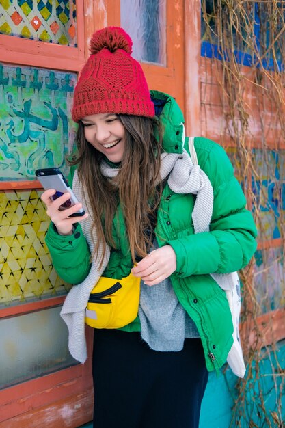 Mujer joven feliz con un gorro de punto rojo, chaqueta verde, bufanda gris, bolso pequeño amarillo. Hipster chica en ropa brillante sosteniendo un teléfono inteligente en la mano, riendo y sonriendo.