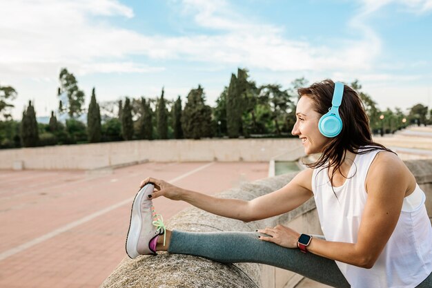 Foto mujer joven feliz haciendo ejercicio de estiramiento de las piernas antes de correr mientras escucha música al aire libre
