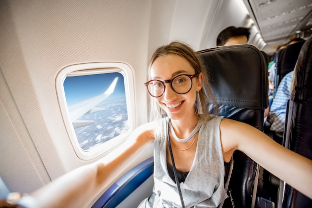 Mujer joven feliz haciendo foto selfie sentado en el asiento del avión cerca de la ventana durante el vuelo en el avión