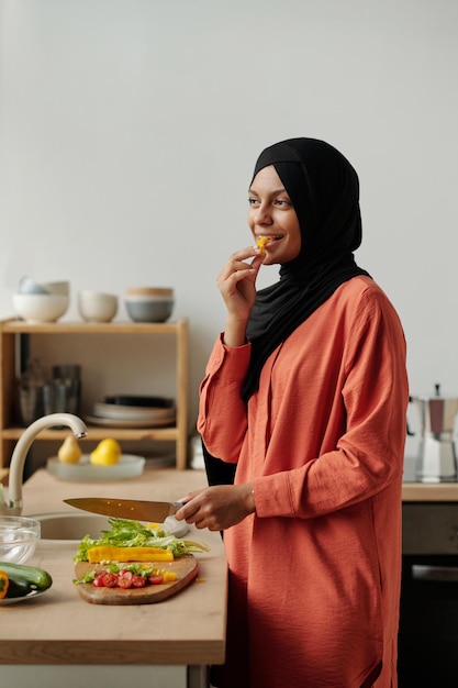 Mujer joven feliz con hijab y camisa roja comiendo una rebanada de pimienta amarilla fresca