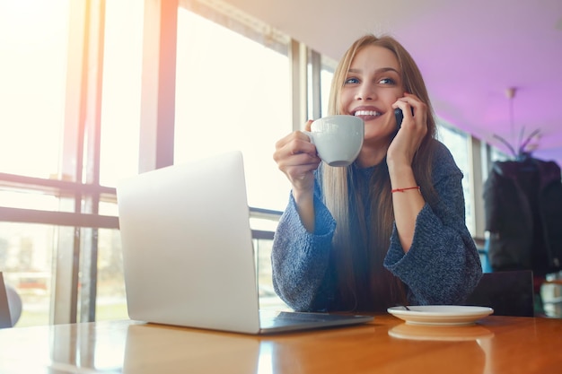 Mujer joven feliz hipster con taza de café hablando por teléfono en la computadora portátil del café en la mesa