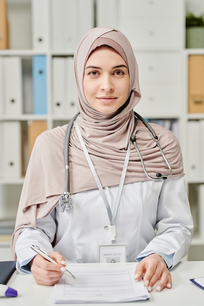 Mujer joven feliz en hiyab y bata blanca tomando notas en un documento médico