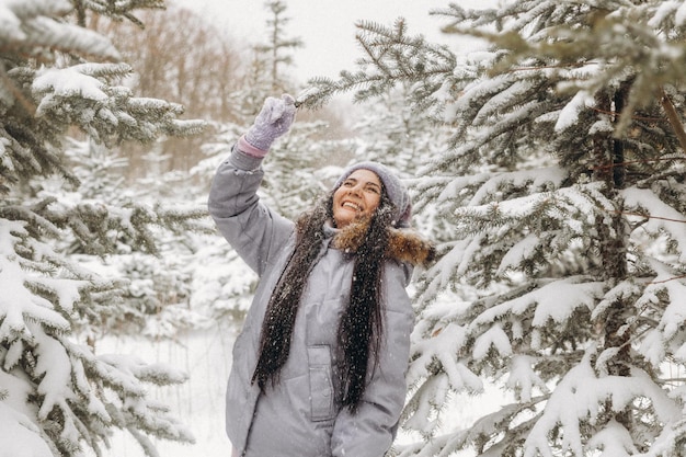 Mujer joven feliz en invierno en un paseo por la naturaleza. una mujer con una chaqueta lila se encuentra cerca de los árboles de Navidad en un parque de invierno.