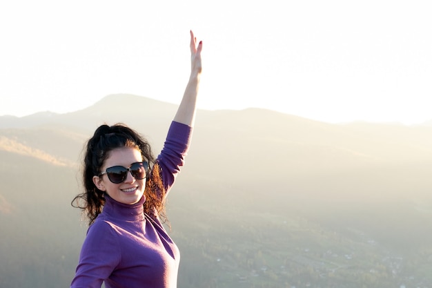 Mujer joven feliz con la mano levantada disfrutando de la cálida tarde del atardecer en las montañas de verano.
