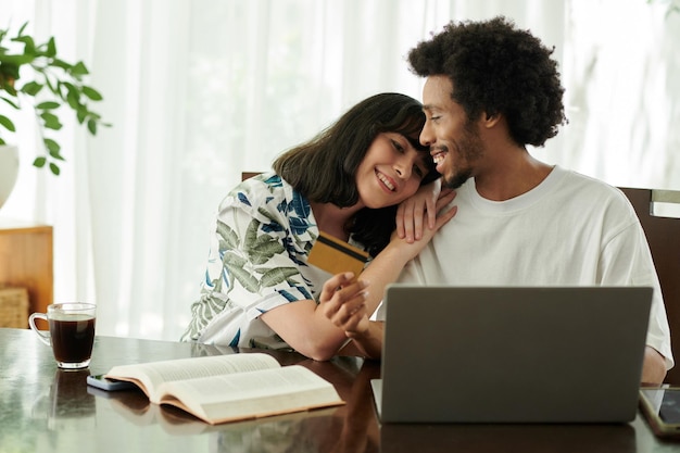 Mujer joven feliz manteniendo la cabeza sobre el hombro de su marido con tarjeta de crédito mientras ambos están sentados frente a la computadora portátil y ordenando productos en línea