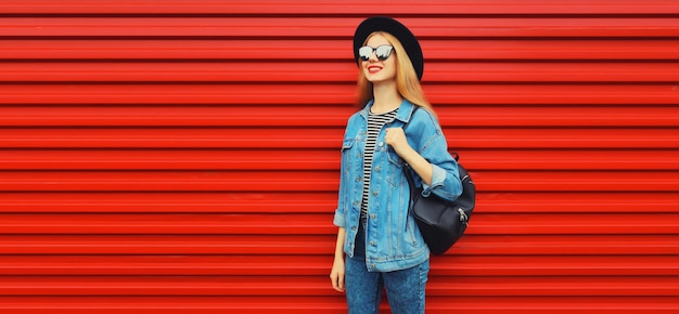 mujer joven feliz mirando lejos en negro sombrero redondo chaqueta de jean mochila en fondo rojo