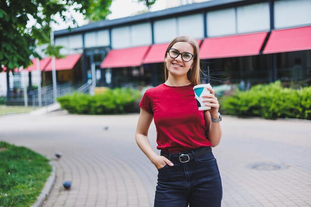 Mujer joven feliz con mochila sonriendo y mirando hacia otro lado mientras camina por la calle de la ciudad y disfruta de un café recién hecho para llevar