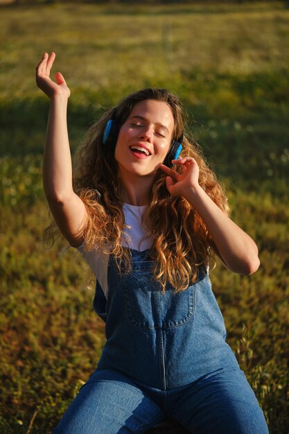 Mujer joven feliz en mono de vaqueros bailando con el brazo levantado y cantando mientras escucha buena música en auriculares y sentada con los ojos cerrados en el prado cubierto de hierba en el campo