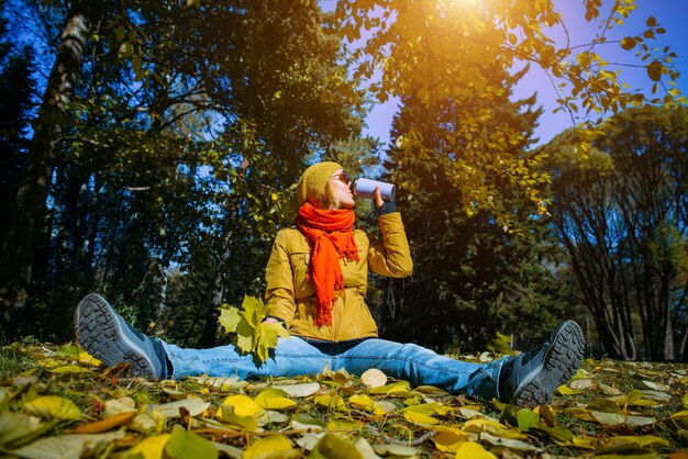 Mujer joven feliz en el parque de otoño en un día soleado, tomando café o té, sentado sobre el follaje que cae, divirtiéndose con hojas amarillas. Concepto de estilo de vida positivo.