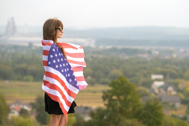 Mujer joven feliz con el pelo largo sosteniendo ondeando en el viento la bandera nacional americana en sus hombros descansando al aire libre disfrutando de un cálido día de verano.