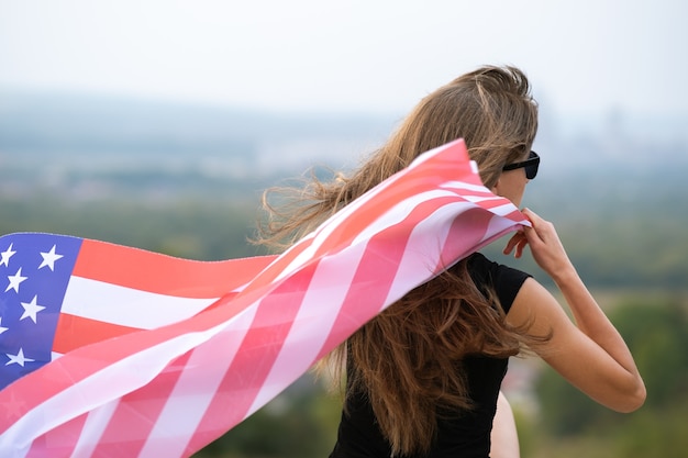 Mujer joven feliz con el pelo largo sosteniendo ondeando en el viento de la bandera nacional americana