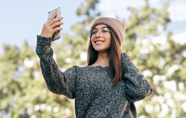 Mujer joven feliz de pie al aire libre contra la pared del edificio y tomando autorretrato en el teléfono inteligente Hermosa mujer vestida con suéter gris sombrero rosa anteojos transparentes haciendo selfie afuera