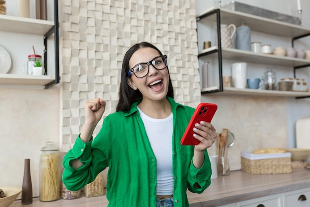 Mujer joven feliz de pie en la cocina en casa y mirando a la cámara usa un teléfono móvil