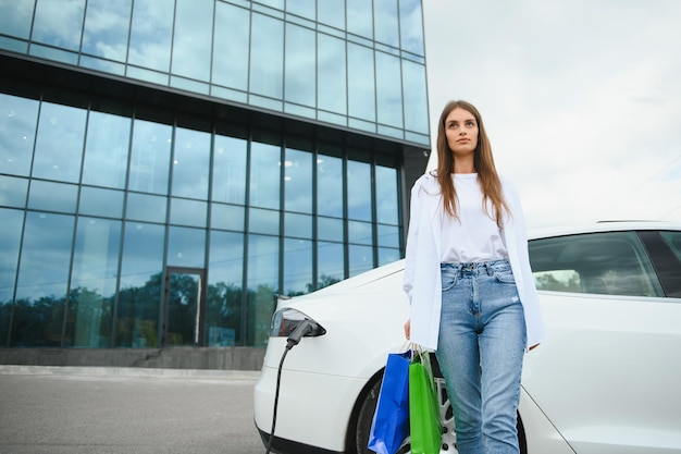 Mujer joven feliz de pie en el estacionamiento de la ciudad cerca de un coche eléctrico que carga la batería del automóvil desde una pequeña estación de la ciudad con bolsas de compras