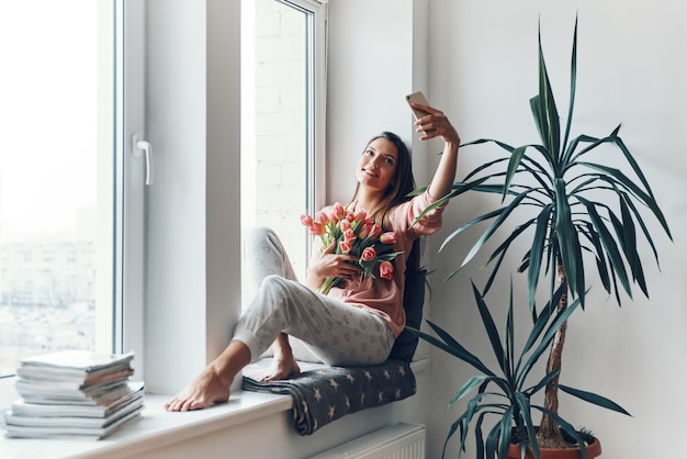 Mujer joven feliz en pijama acogedor tomando selfie con su ramo de tulipanes mientras descansa en el alféizar de la ventana en casa