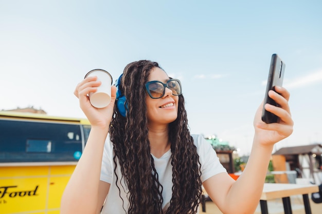 Una mujer joven feliz que asiste a una conferencia web a través de auriculares o un trabajador independiente sentado en una mesa de café al aire libre una chica con rastas se comunica a través de un enlace de video