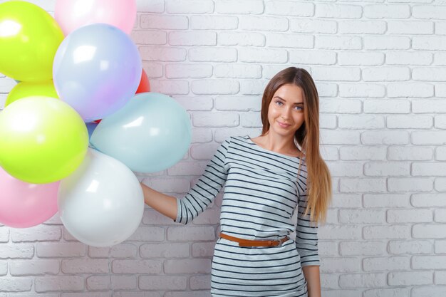 Mujer joven feliz que se coloca sobre la pared de ladrillo y que sostiene los globos.