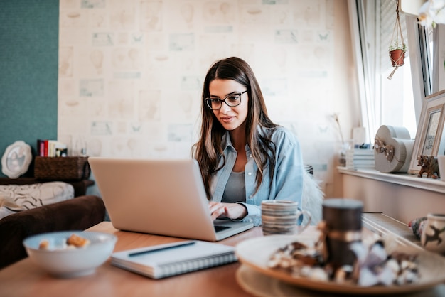 Mujer joven feliz que estudia en la computadora portátil en su apartamento.