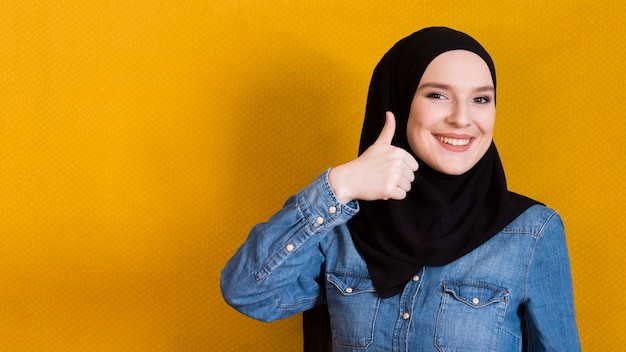 Mujer joven feliz que gesticula el thumbup contra superficie amarilla brillante